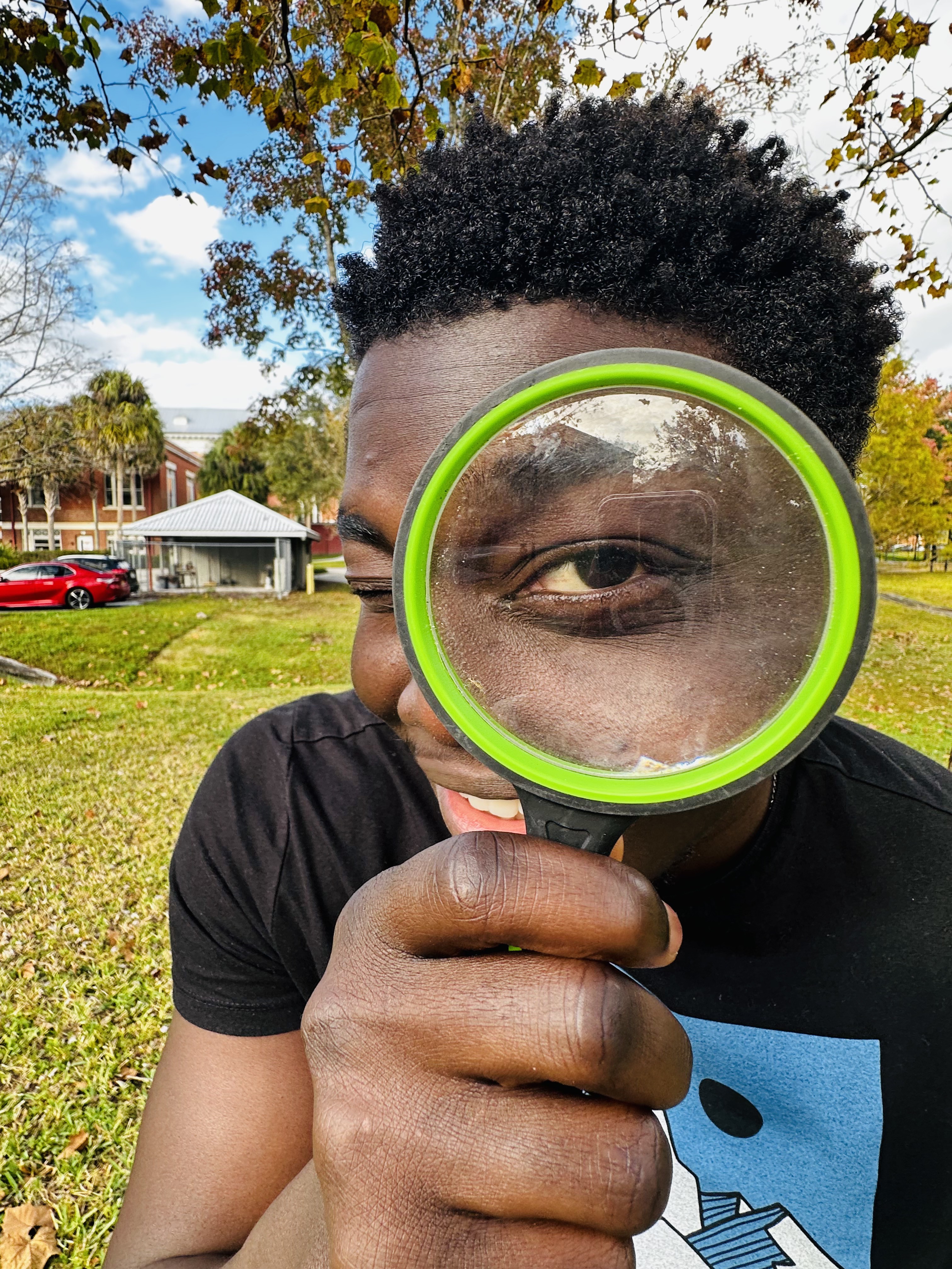Simon outdoors, smiling with a green magnifying glass held over one eye